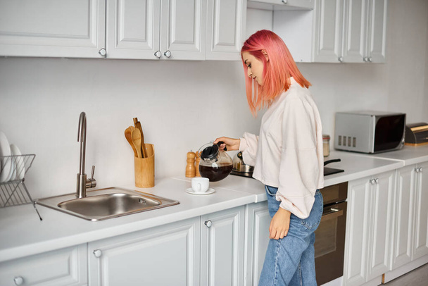 dobrze wyglądająca młoda kobieta w stroju codziennym z różowymi włosami nalewająca sobie kawy podczas pobytu w kuchni - Zdjęcie, obraz