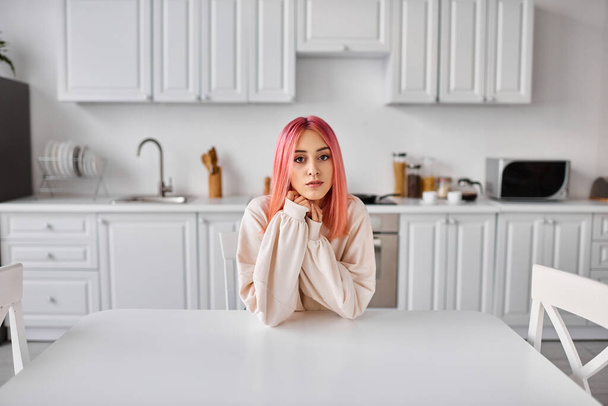 piękna młoda kobieta z różowymi włosami siedzi w kuchni przy stole i patrzy prosto w kamerę - Zdjęcie, obraz