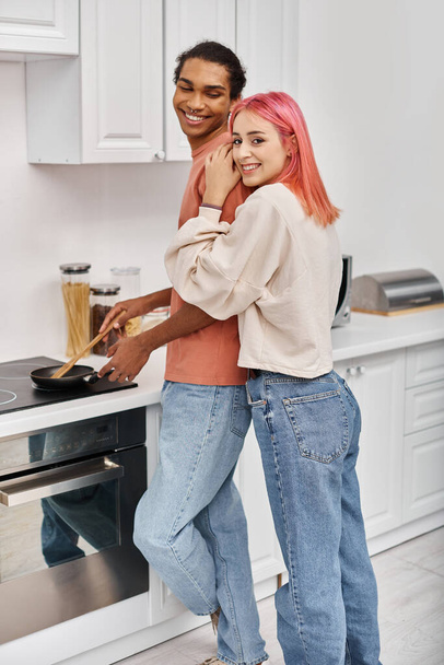 gaie femme aux cheveux roses en vêtements de maison étreignant son petit ami afro-américain qui cuisine le dîner - Photo, image