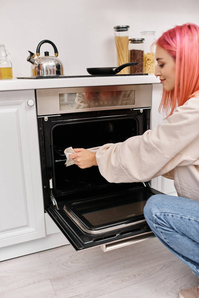 молода приваблива жінка з рожевим волоссям в повсякденному затишному одязі, використовуючи піч, щоб приготувати вечерю вдома - Фото, зображення