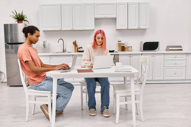 appealing hard working interracial couple sitting at table in kitchen with laptops, freelance - Photo, Image