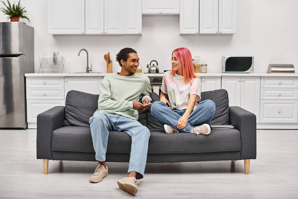 atraente casal alegre diverso sentado no sofá na sala de estar e sorrindo um para o outro em casa - Foto, Imagem