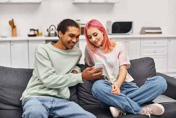 fröhliches multiethnisches Paar in lässiger Kleidung auf dem Sofa sitzend und zu Hause auf das Smartphone schauend - Foto, Bild