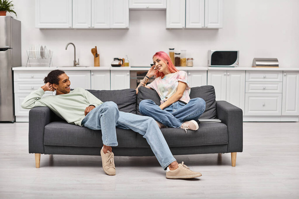 good looking joyous interracial couple relaxing on sofa in living room and smiling at each other - Photo, Image