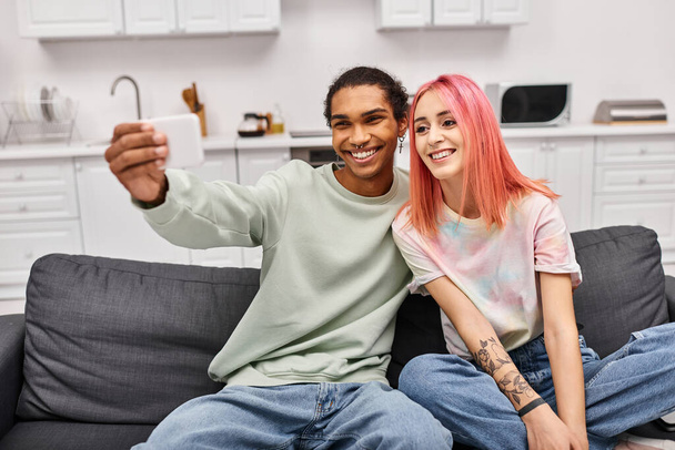 fröhlich attraktives multikulturelles Paar in Hauskleidung, das Selfies macht, während es zu Hause im Wohnzimmer ist - Foto, Bild