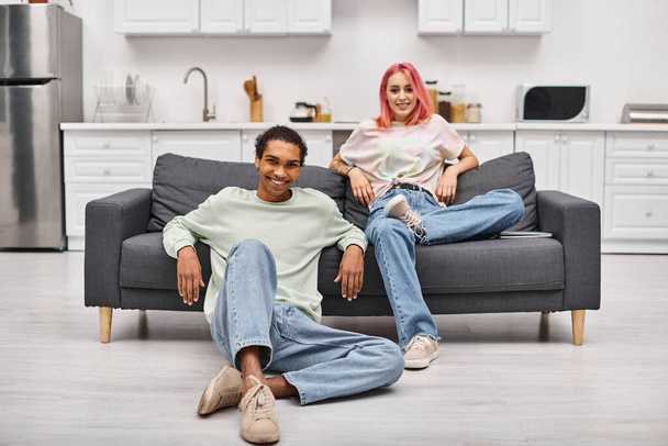 joyous attractive multiracial couple in cozy homewear posing in living room and smiling at camera - Photo, Image