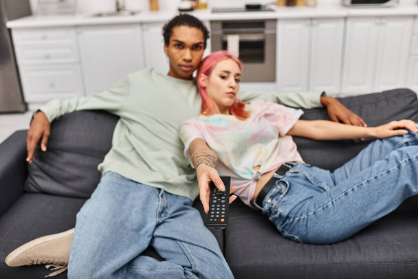 focus on remote control in hand of pretty blurred woman and her african american boyfriend at home - Photo, Image