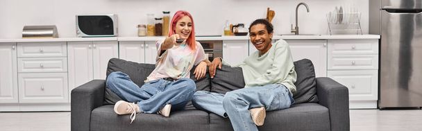 radostný mladý mezirasový pár v pohodlném oblečení sedí na pohovce a sleduje filmy, banner - Fotografie, Obrázek