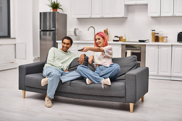 iloinen houkutteleva monipuolinen pari viihtyisissä asuissa istuu sohvalla ja katselee elokuvia yhdessä - Valokuva, kuva