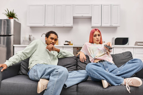 verängstigtes multirassisches Paar in lässiger Kleidung, das zu Hause im Wohnzimmer Horrorfilme anschaut - Foto, Bild