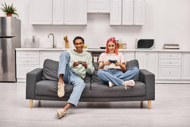 heureux couple interracial dans les vêtements de maison assis sur le canapé et jouer à des jeux en utilisant des manettes de jeu à la maison - Photo, image