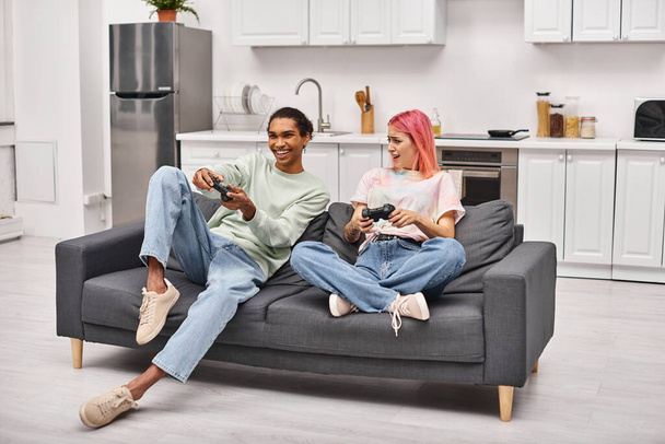 couple multiculturel joyeux dans des vêtements confortables assis et jouer à des jeux vidéo avec joysticks à la maison - Photo, image
