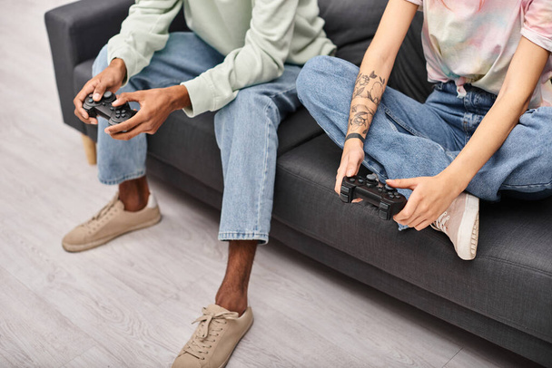 abgeschnittene Ansicht eines jungen multikulturellen Paares in lässiger Kleidung, das zu Hause Spiele mit Gamepads spielt - Foto, Bild