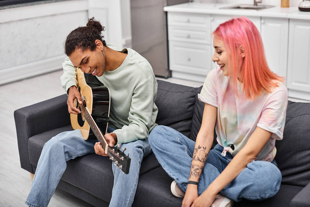 jolly pink haired woman watching her handsome cheerful boyfriend playing guitar while at home - Photo, Image