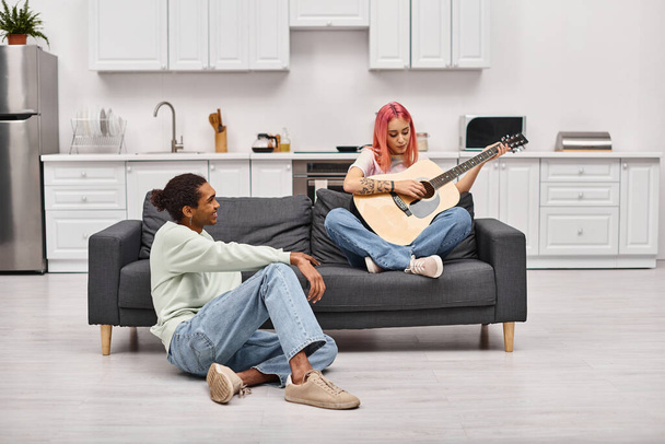 homme afro-américain joyeux en vêtements de maison regardant avec amour sa petite amie aux cheveux roses jouer de la guitare - Photo, image