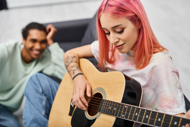 enfoque en la mujer de pelo rosa tocando la guitarra delante de su novio afroamericano alegre borrosa - Foto, Imagen