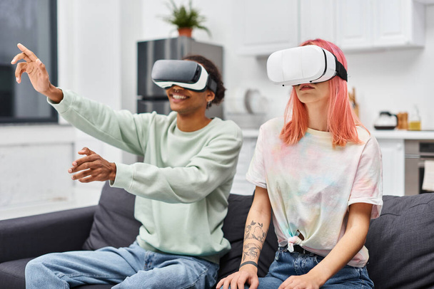radosna, atrakcyjna, różnorodna para w luźnym stroju, siedząca na kanapie w salonie z zestawami słuchawkowymi VR - Zdjęcie, obraz
