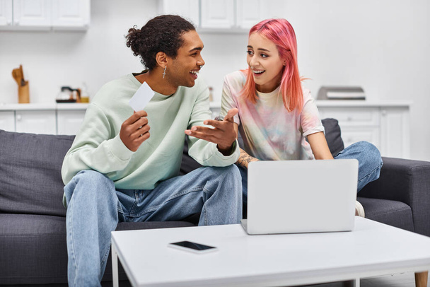 vreugdevolle interraciale paar in homewear houden credit card en glimlachen naar elkaar in de buurt laptop - Foto, afbeelding