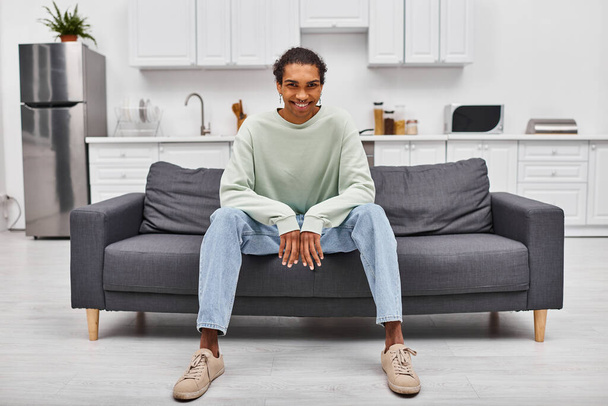Χαρούμενος νεαρός Αφροαμερικάνος με άνετο ένδυμα κάθεται στον καναπέ και κοιτάζει την κάμερα στο σπίτι - Φωτογραφία, εικόνα