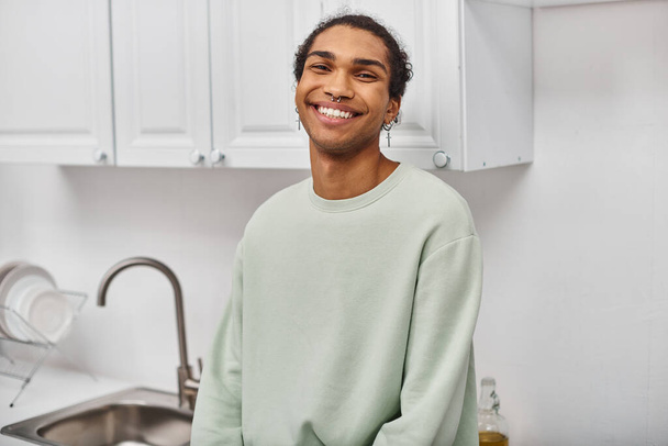wesoły, atrakcyjny, afrykański Amerykanin w białym ciepłym swetrze uśmiechający się szczęśliwie do kamery w domu - Zdjęcie, obraz