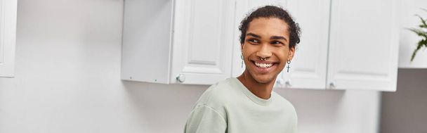 хорошо выглядящий жизнерадостный африканский мужчина в зеленом свитере, отводящий взгляд, находясь на кухне, баннер - Фото, изображение