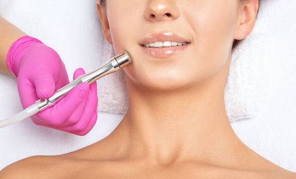 Der Kosmetologe führt das Verfahren Mikrodermabrasion der Gesichtshaut und des Halses eines schönen Mädchens in einem Schönheitssalon durch. Kosmetologie und professionelle Hautpflege. - Foto, Bild