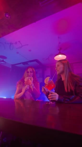 Две счастливые женщины разговаривают в ночном баре в неоновом освещении и пьют алкогольные коктейли. Концепция ночного клуба и женской дружбы. Медленное движение. Вертикальное видео. - Кадры, видео