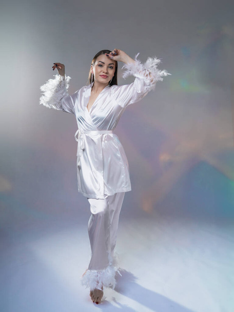 Hermosa mujer en pijama blanco satinado con plumas. Morena sobre un fondo claro - Foto, Imagen