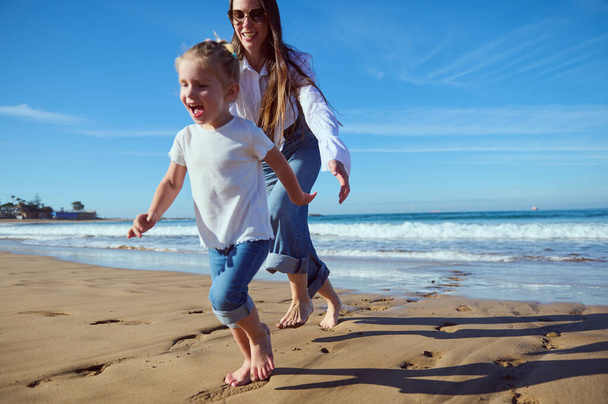 Retrato de comprimento total de uma família feliz de uma menina pequena ativa bonita caucasiana e sua mãe rindo, sorrindo, brincando juntos e correndo descalços na praia - Foto, Imagem