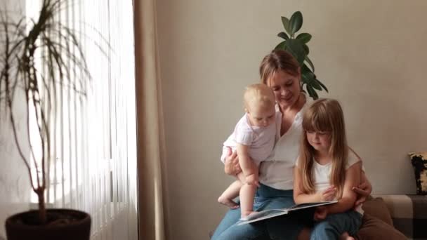 Mom read book for her little daughters on sofa at home. Preschool girl enjoy time and fascinating fairytale rest with her younger baby sister and mother. Kids development, intellectual growth concept - Footage, Video