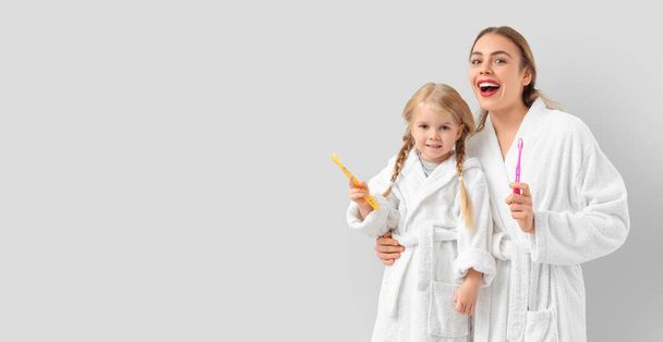 Young mother and her little daughter in bathrobes and with toothbrushes on light background with space for text - Photo, Image