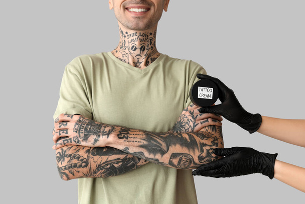 Maestro del tatuaje en guantes sosteniendo tarro de crema cerca del hombre con tatuajes sobre fondo gris - Foto, imagen