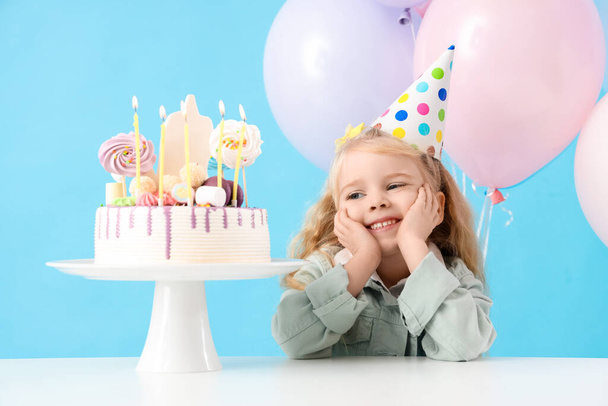 Schattig klein meisje in feestmuts met ballonnen en verjaardagstaart op tafel tegen een blauwe achtergrond - Foto, afbeelding