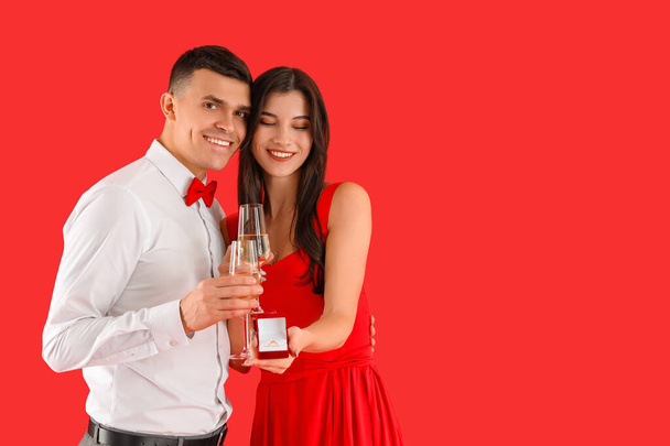 Молодая обрученная пара с обручальным кольцом и бокалами шампанского на красном фоне. Празднование Дня Святого Валентина - Фото, изображение
