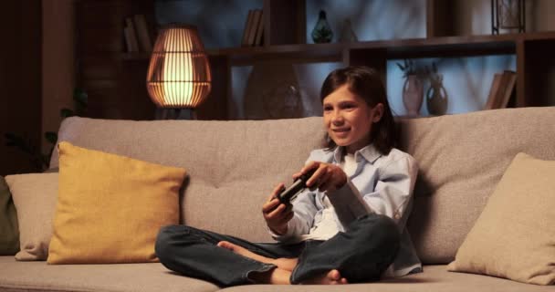Zdeterminowany chłopiec siedzi na kanapie, poruszając się po grze wideo z gamepadem w ręku. Jego oczy są przyklejone do ekranu, a palce poruszają się umiejętnie po przyciskach.. - Materiał filmowy, wideo