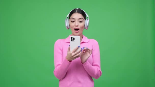 Caucasian woman, wearing headphones and holding a phone, is surprised by good news, creating a visually engaging scene filled with positive emotions and genuine delight over green background. - Footage, Video