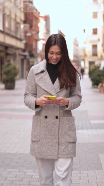 Молодая латиноамериканка пользуется мобильным телефоном. Она ходит и улыбается на улице. Высококачественные кадры - Кадры, видео