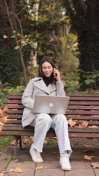 Młoda kobieta pracująca w laptopie i mówiąca w smartfonie w parku. Pochodzenie łacińskie. Materiał wysokiej jakości - Materiał filmowy, wideo