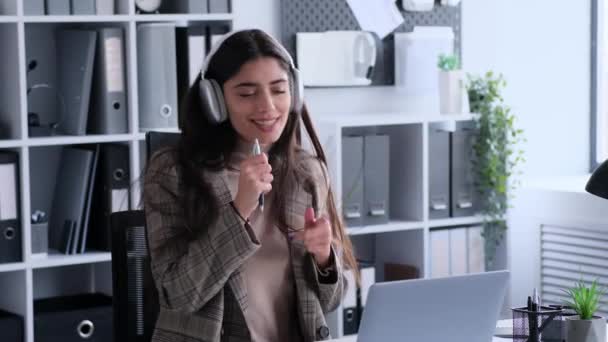 Een vrouw die positiviteit uitstraalt, luistert naar muziek in een koptelefoon en zingt vrolijk tijdens het werk op kantoor. Ze straalt verheffende en energieke ambiance uit die ze brengt naar professionele taken. - Video