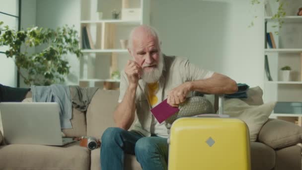 Ein enttäuschter alter Mann mit Reisepass und Koffer. Ein älterer Mann ist unglücklich über einen missglückten Trip und sitzt auf dem Sofa. Passprobleme. Reiseplanung. - Filmmaterial, Video