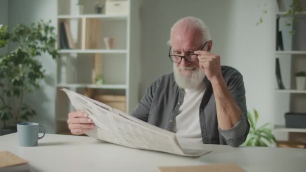 Un vecchio sorridente e intelligente siede a un tavolo e legge un giornale.Un uomo anziano con un giornale ride, vede le notizie e gli eventi attuali nel giornale, Concetto quotidiano - Filmati, video