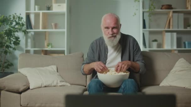 Vanha mies syö popcornia ja katselee urheiluohjelmaa.Vakava iäkäs mies istuu sohvalla ja juhlii urheiluvoittoa. Elokuvat rentoutumisen ja psykologisen helpotuksen muotona. - Materiaali, video