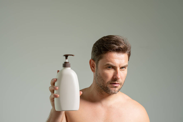 Koncept kosmetických procedur péče o pleť. mladý pohledný muž usmívá s veselým, sebevědomým výrazem s rukou na bradě s županem a drží láhev šamponu připravující se na mytí - Fotografie, Obrázek