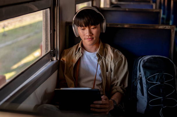 Een gelukkige en ontspannen jonge Aziatische mannelijke reiziger luistert naar muziek op zijn koptelefoon en gebruikt zijn digitale tablet tijdens het woon-werkverkeer met de trein naar ergens. solo reizen, backpacker, vakantie - Foto, afbeelding