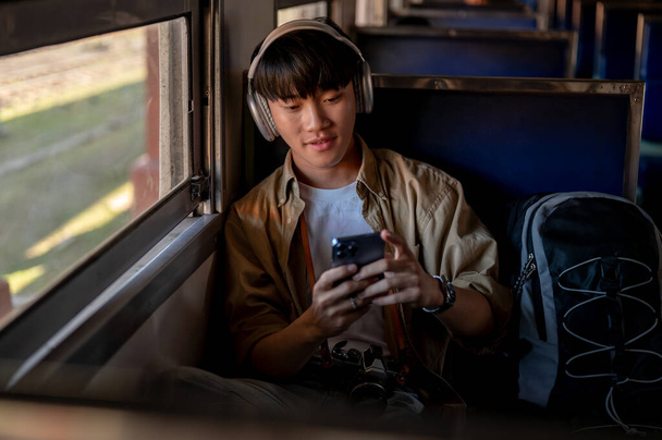 Een vrolijke en ontspannen jonge Aziatische mannelijke reiziger luistert naar muziek op zijn koptelefoon en gebruikt zijn smartphone tijdens het woon-werkverkeer met de trein naar ergens. solo reizen, backpacker, vakantie - Foto, afbeelding