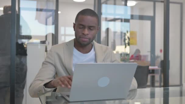 africano uomo d'affari sorridente a fotocamera mentre si lavora su computer portatile in ufficio - Filmati, video