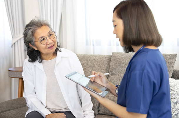 Μια ηλικιωμένη γυναίκα που δίνει μια συνέντευξη για την υγεία σε ένα σπίτι επίσκεψη νοσοκόμα στο σπίτι, μια νεαρή γυναίκα εμπειρογνωμοσύνη στον τομέα της υγειονομικής περίθαλψης μιλάμε και τη συλλογή δεδομένων σε tablet υπολογιστή, έννοια της κατ 'οίκον περίθαλψης, κατ' οίκον υγειονομική περίθαλψη - Φωτογραφία, εικόνα
