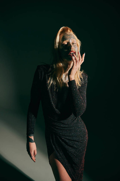 татуйована блондинка з блиском на обличчі і червоними губами позує в блискучій сукні на чорному тлі - Фото, зображення