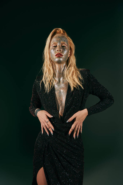 татуированная блондинка с блестками на лице и теле позирует в блестящем платье на черном фоне - Фото, изображение