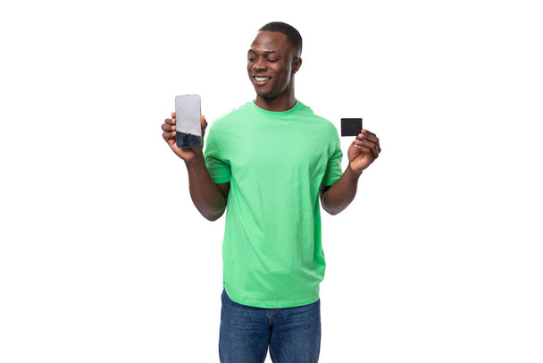 młody amerykański facet ubrany w miętowy t-shirt z smartfonem i kartą kredytową. - Zdjęcie, obraz